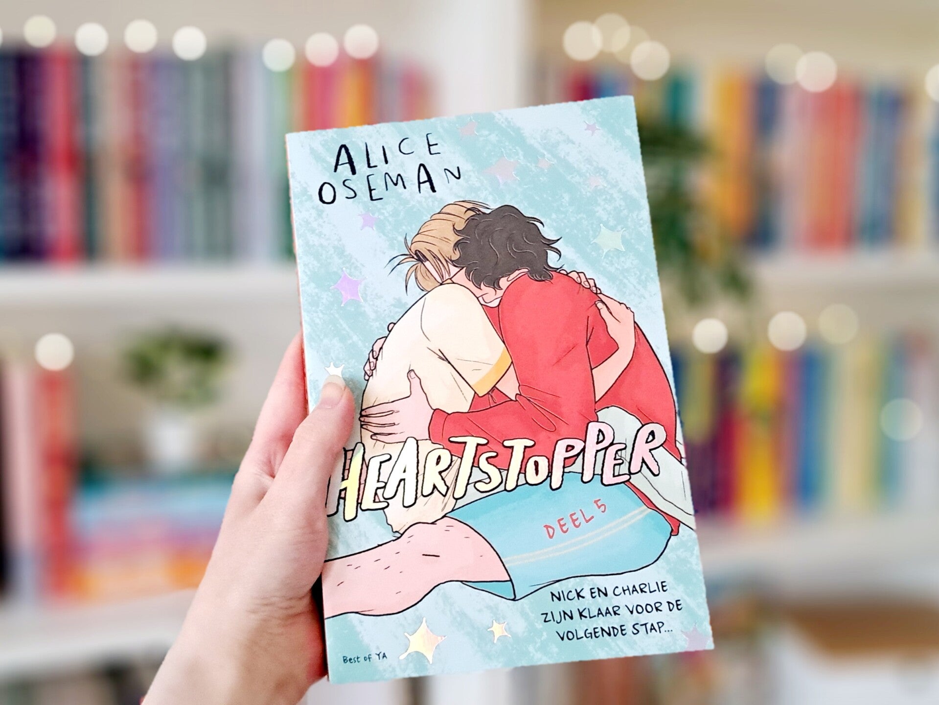Een witte hand houdt 'Heartstopper deel 5: nick en charlie zijn klaar voor de volgende statp' van Alice Oseman omhoog voor een kleurrijke wazige boekenkast met lichtjes. Foto bij recensie door turnthepages.nl