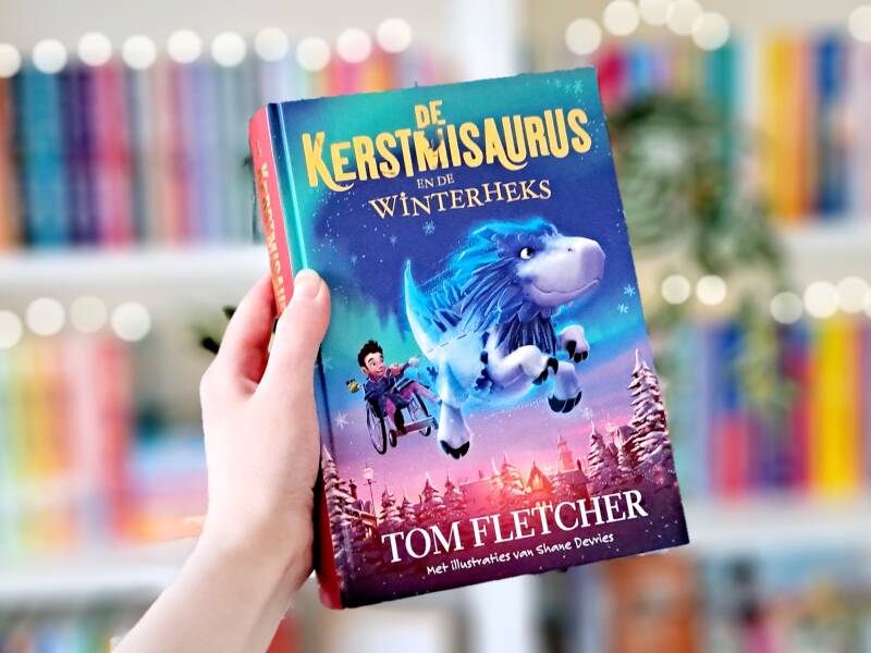 Een witte hand houdt 'De kerstmisaurus en de winterheks' van Tom Fletcher omhoog voor een kleurrijke wazige boekenksat met lichtjes. Foto bij own-voices recensie door turnthepages.nl Rolstoelgebruiker en disabled bookstagrammer en blogger.
