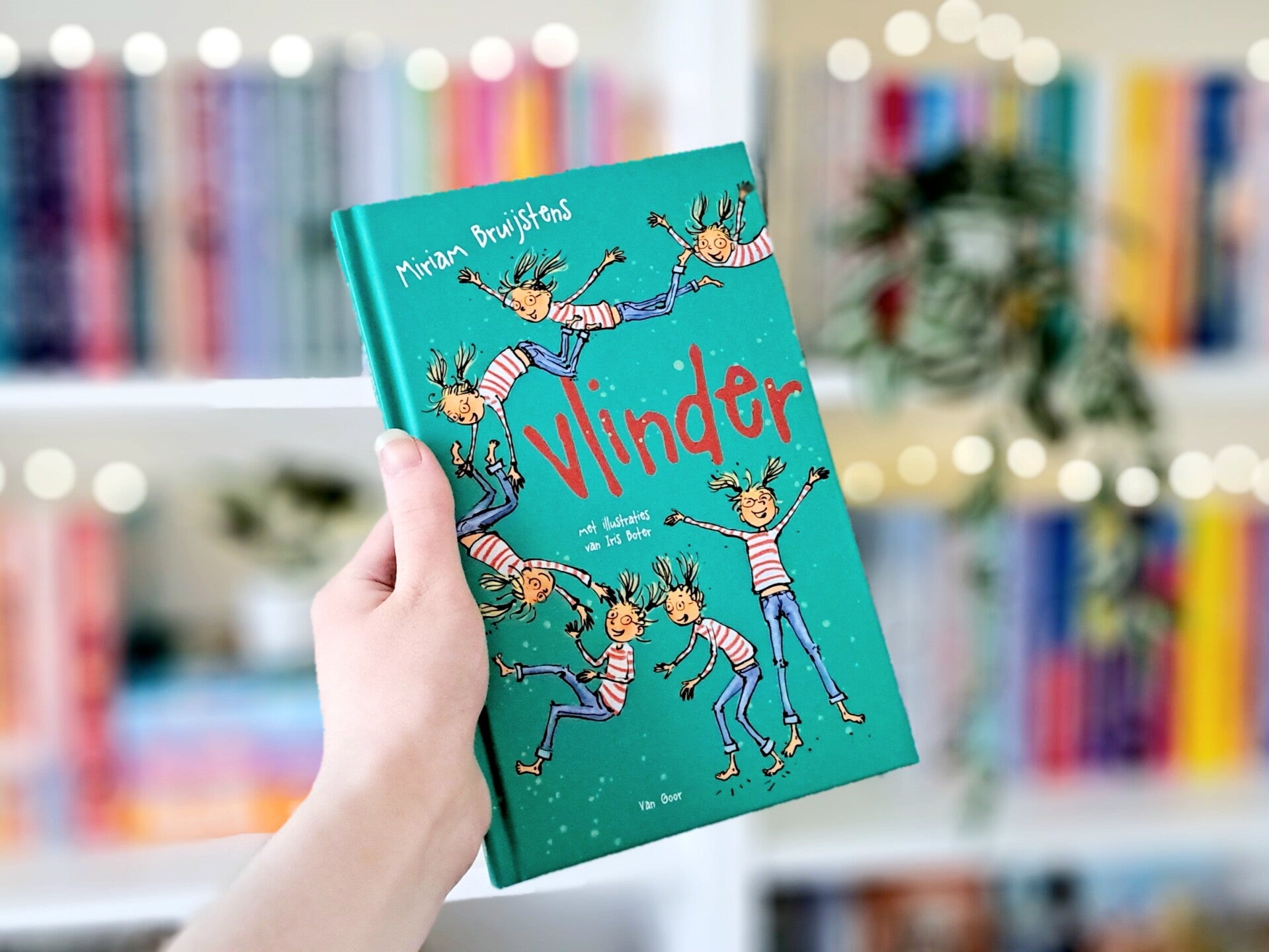 Een witte hand houdt 'Vlinder' van Miriam Bruijstens omhoog voor een kleurrijke wazige boekenkast met lichtjes. Foto bij own-voices recensie door turnthepages.nl | Disabled bookstagrammer en boekblogger met ADHD.