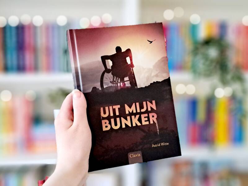 Een witte hand houdt 'Voorbij het wederwoud' van Valentijn Ringelberg omhoog voor een kleurrijke wazige boekenkast met lichtjes. Foto bij own-voices recensie door turnthepages.nl Disabled bookstagrammer en blogger.