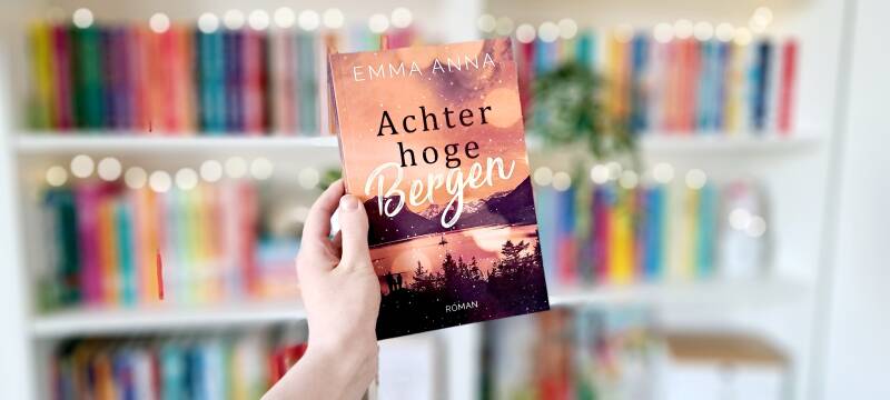 Een witte hand houdt 'Achter hoge bergen' van Emma Anna omhoog voor een kleurrijke wazige boekenkast met lichtjes. Foto bij recensie door turnthepages.nl Rolstoelgebruiker, disabled en chronisch ziek bookstagrammer en boekblogger.