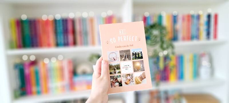 Een witte hand houdt '(Not) so perfect' van Linda van de Pol omhoog voor een kleurrijke wazige boekenkast met lichtjes. Foto bij recensie door turnthepages.nl Disabled bookstagrammer en boekblogger.