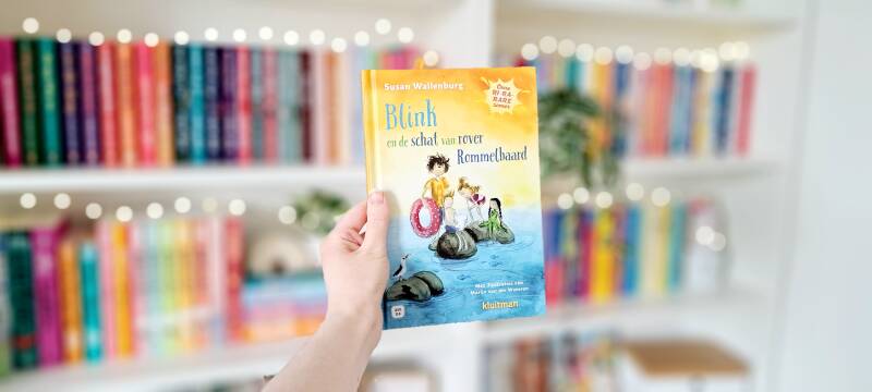 Een witte hand houdt 'Blink en de schat van rover Rommelbaard', deel 1 in de serie Onze Ri-ra-rare zomer van Susan Wallenburg. geïllustreerd door Marijn van der Wateren omhoog voor een kleurrijke wazige boekenkast met lichtjes. Foto bij own-voices recensi