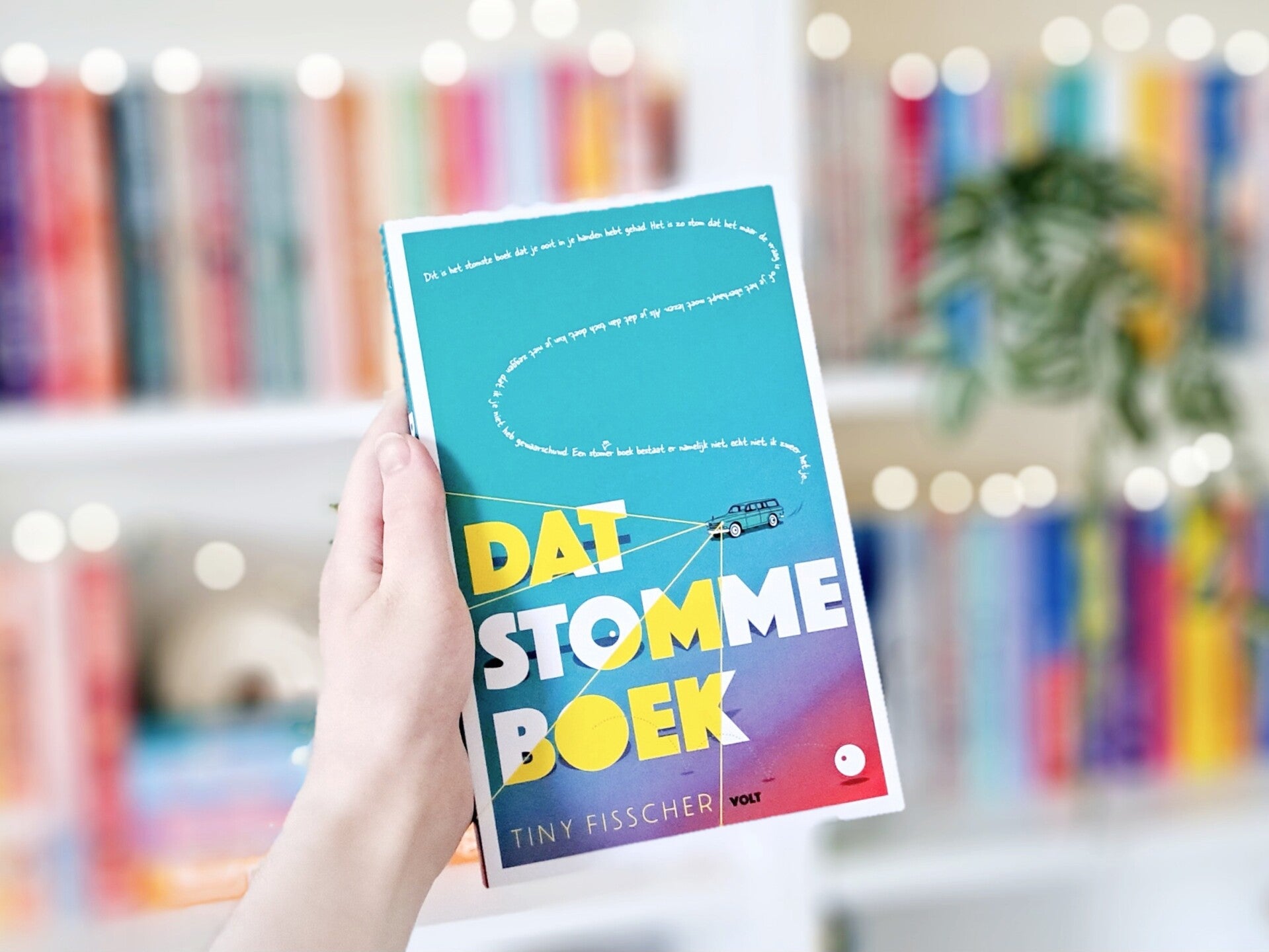 Ee witte hand houdt 'Dat stomme boek' van Tiny Fisscher omhoog voor een kleurrijke wazige boekenkast met lichtjes. Foto bij recensie door turnthepages.nl Disabled bookstagrammer en boekblogger met ADHD.