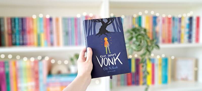 Een witte hand houdt 'een soort vonk' van Elle McNicoll omhoog voor een kleurrijke wazige boekenkast met lichtjes. Recensie door Annemarie van turnthepages.nl | Disabled bookstagrammer en neurodivergent boekblogger.