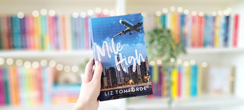 Een witte hand houdt 'Mile High' het eerste deel in de Windy City serie van Liz Tomforde omhoog voor een kleurrijke wazige boekenkast met lichtjes. Foto bij recensie door turnthepages. Disabled bookstagrammer en boekblogger.