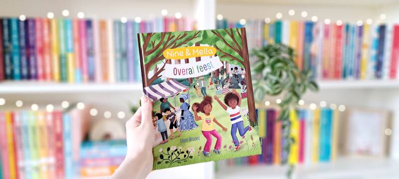 Een witte hand houdt 'Nine en Mella - Overal feest' geschreven door Ellen Brudet en geïllustreerd door Maruga Koops omhoog voor een kleurrijke wazige boekenkast met lichtjes. Foto bij own-voices recensie door turnthepages.nl Rolstoelgebruiker en disabled 
