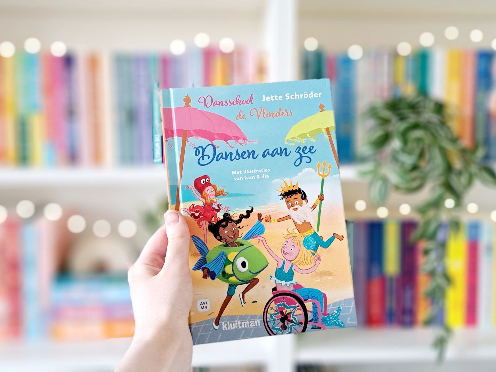 Een witte hand houdt 'Dansen aan zee' deel twee in AVI serie Dansschool de Vlinders van Jette Schröder, geïllustreerd door ivan en ilia, omhoog voor een kleurrijke wazige boekenkast met lichtjes. Foto bij own-voices recensie door turnthepages.nl | Rolstoe