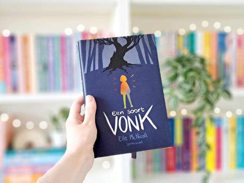 Een witte hand houdt 'Een soort vonk' van Elle McNicoll omhoog voor een kleurrijke wazige boekenkast met lichtjes. 