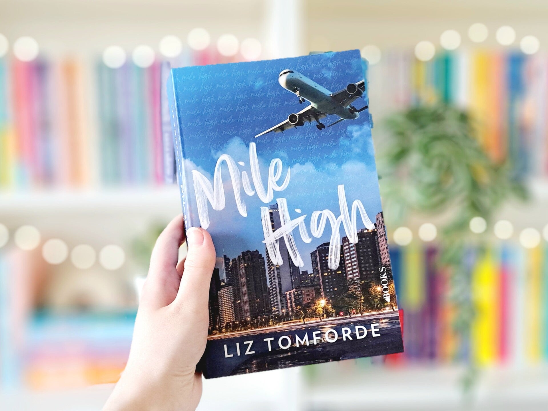 Een witte hand houdt 'Mile High' het eerste deel in de Windy City serie van Liz Tomforde omhoog voor een wazige kleurrijke boekenkast met lichtjes. Foto bij recensie door turnthepages.nl | Disabled bookstagrammer en boekblogger.