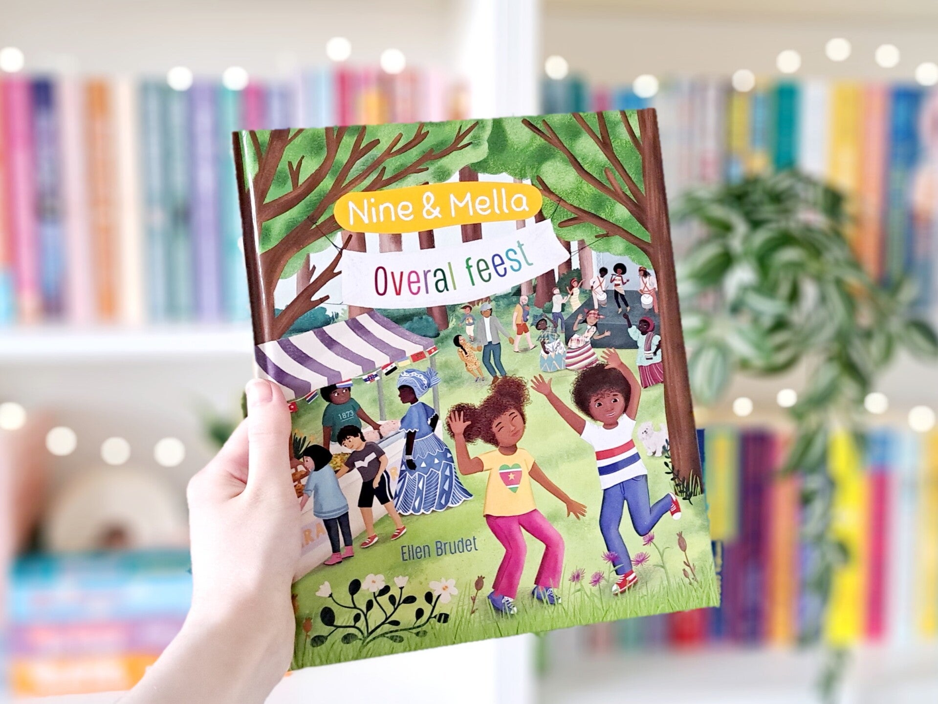 Een witte hand houdt 'Overal feest' van Ellen Brudet en Maruga Koops omhoog voor een kleurrijke wazige boekenkast met lichtjes. Foto bij Own-voices recensie door turnthepages.nl Rolstoelgebruiker en disabled bookstagrammer en blogger met ADHD.