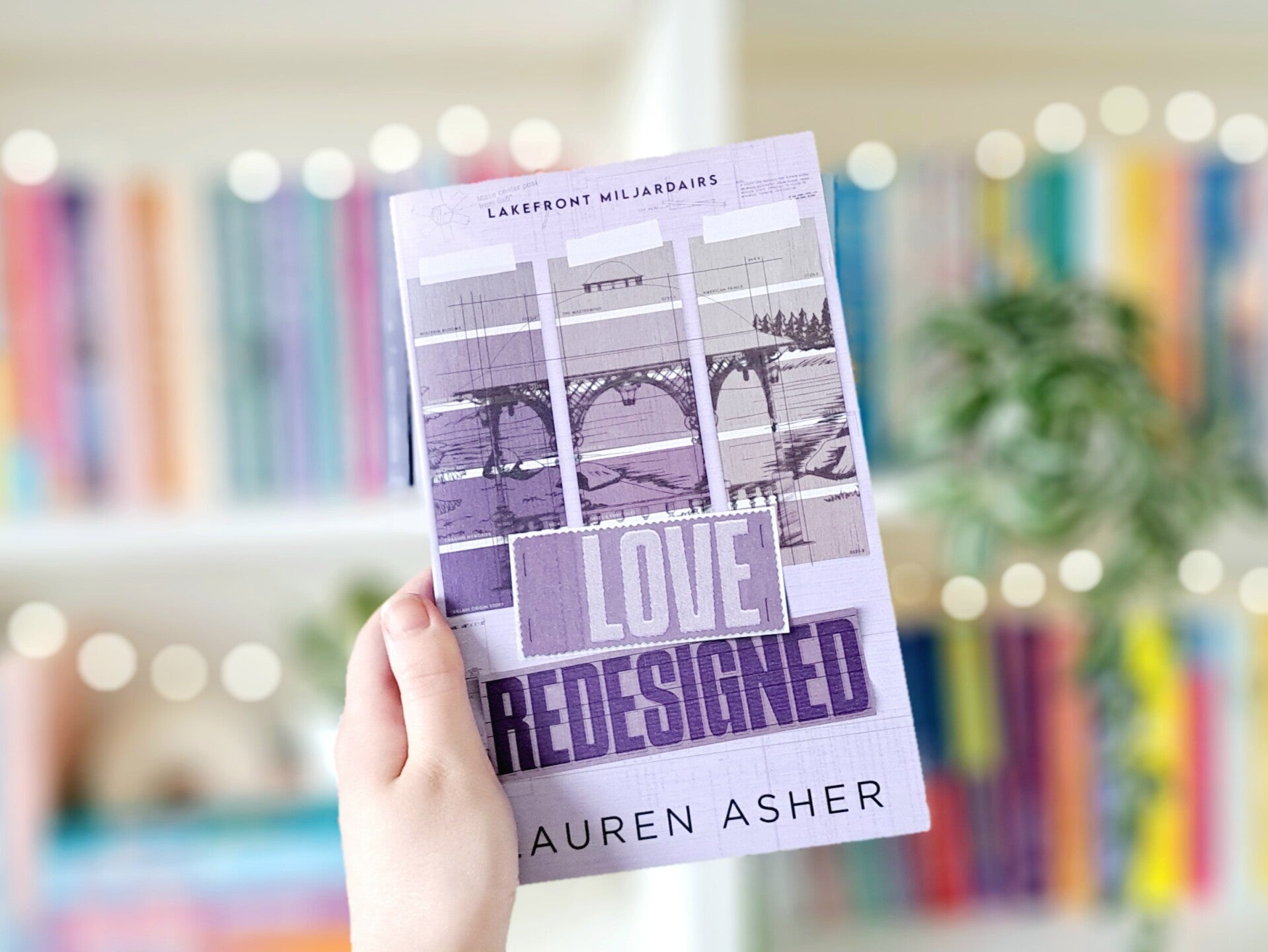 Een witte hand houdt 'Love redesigned' van Lauren Asher omhoog voor een kleurrijke wazige boekenkast met lichtjes. Foto bij recensie door turnthepages.nl | Disabled bookstagrammer en neurodivergente boekblogger.