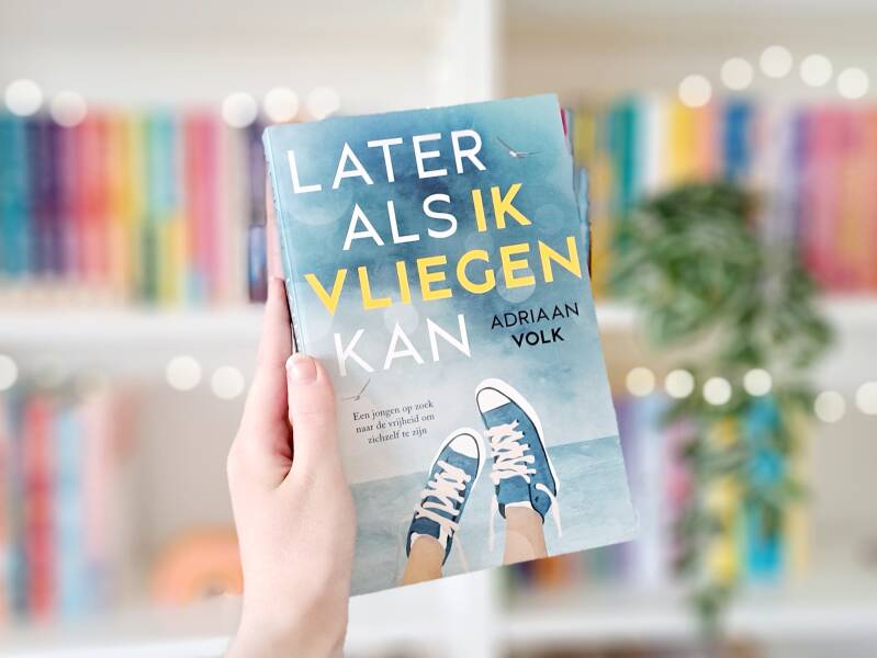 Een witte hand houdt 'Later als ik vliegen kan' van Adriaan VOlk omhoog voor een kleurrijke wazige boekenkast met lichtjes. Recensie door turnthepages.nl Queer en Disabled bookstagramer en boekblogger.