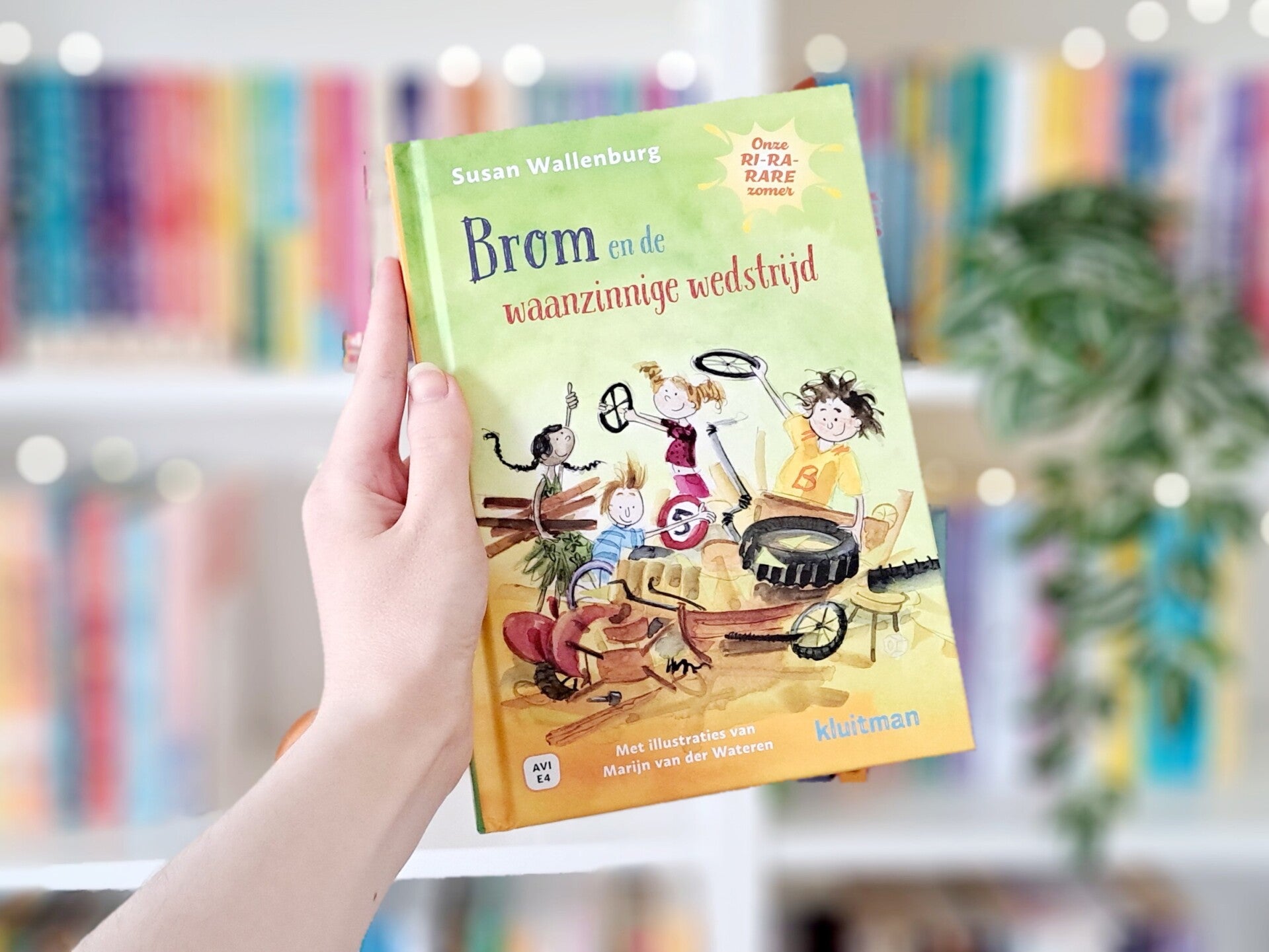 Een witte hand houdt 'Brom en de waanzinnige wedstrijd' van Susan Wallenburg, geillustreerd door Marijn van der Wateren omhoog voor een kleurrijke wazige boekenkast met lichtjes. Foto bij Own-Voices recensie door turnthepages.nl | Rolstoelgebruiker en dis