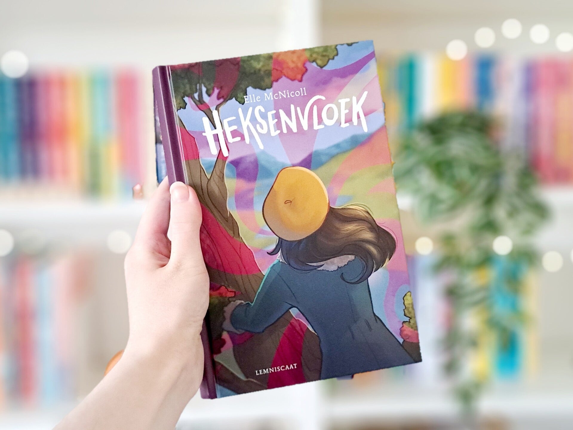 Een witte hand houdt 'Heksenvloek' van Elle McNicoll omhoog voor een kleurrijke wazige boekenkast met lichtjes. Foto bij recensie door turnthepages.nl Rolstoelgebruiker en neurodivergente disabled bookstagrammer en blogger.