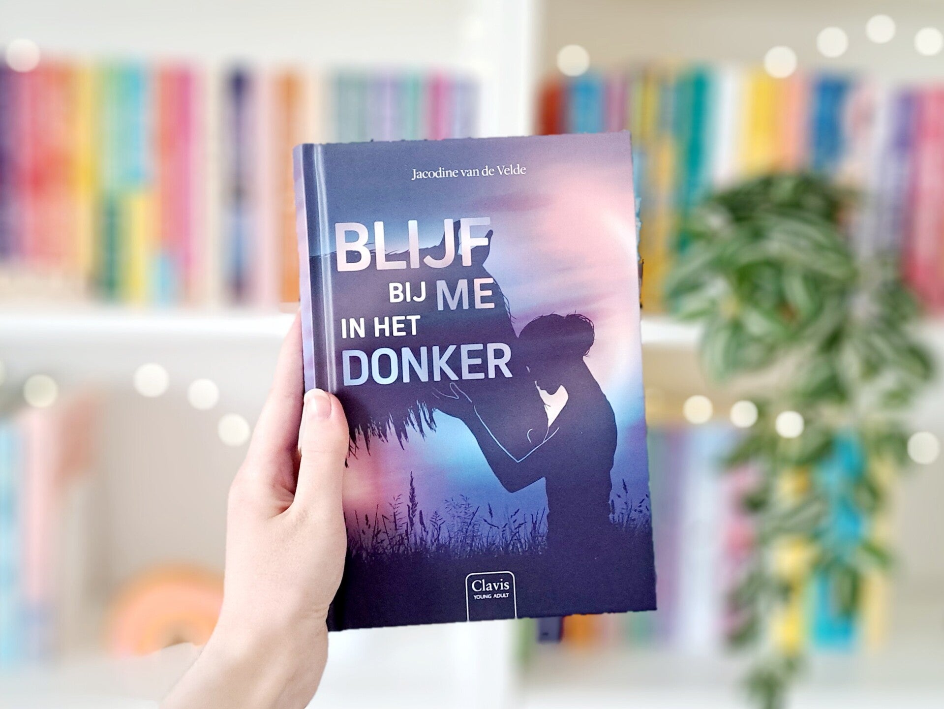 Een witte hand houdt 'Blijf bij me in het donker' van Jacodine van de Velde omhoog voor een kleurrijke wazige boekenkast met lichtjes. Foto bijr ecensie door tunthepages.nl | Disabled bookstagrammer en boekbogger.