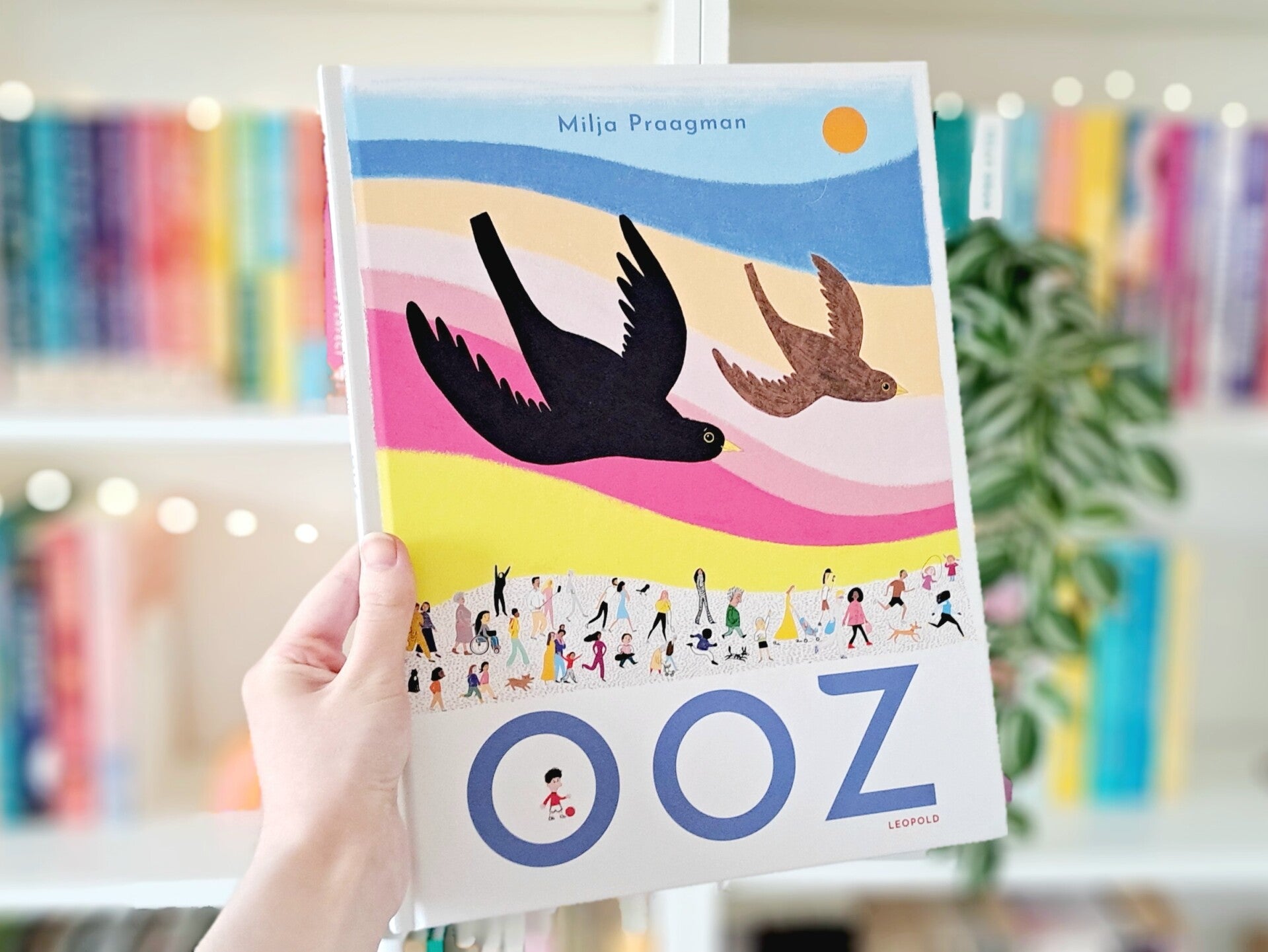 Een witte hand houdt 'Ooz' van Milja Praagman omhoog voor een kleurrijke wazige boekenkast met lichtjes. Foto bij recensie door turnthepages.nl | Rokstoelgebruiker, disabled bookstagrammer en neurodivergente boekblogger.
