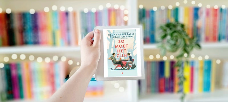 Een witte hand houdt een e-reader met de omslag van 'Zo moet het zijn' van Becky Albertalli en Adam SIlvera omhoog voor een kleurrijke wazige boekenkast met lichtjes. Foto bij own-voices recensie door turnthepages.nl | Disabled bookstagrammer en neurodive
