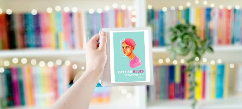 Een witte hand houdt een e-reader met de omslag van 'Catfish blues' van Sara Lövestam omhoog voor een kleurrijke wazige boekenkast met lichtejs. Recensie door turnthepages.nl | Disabled bookstagrammer en neurodivergente boekblogger.