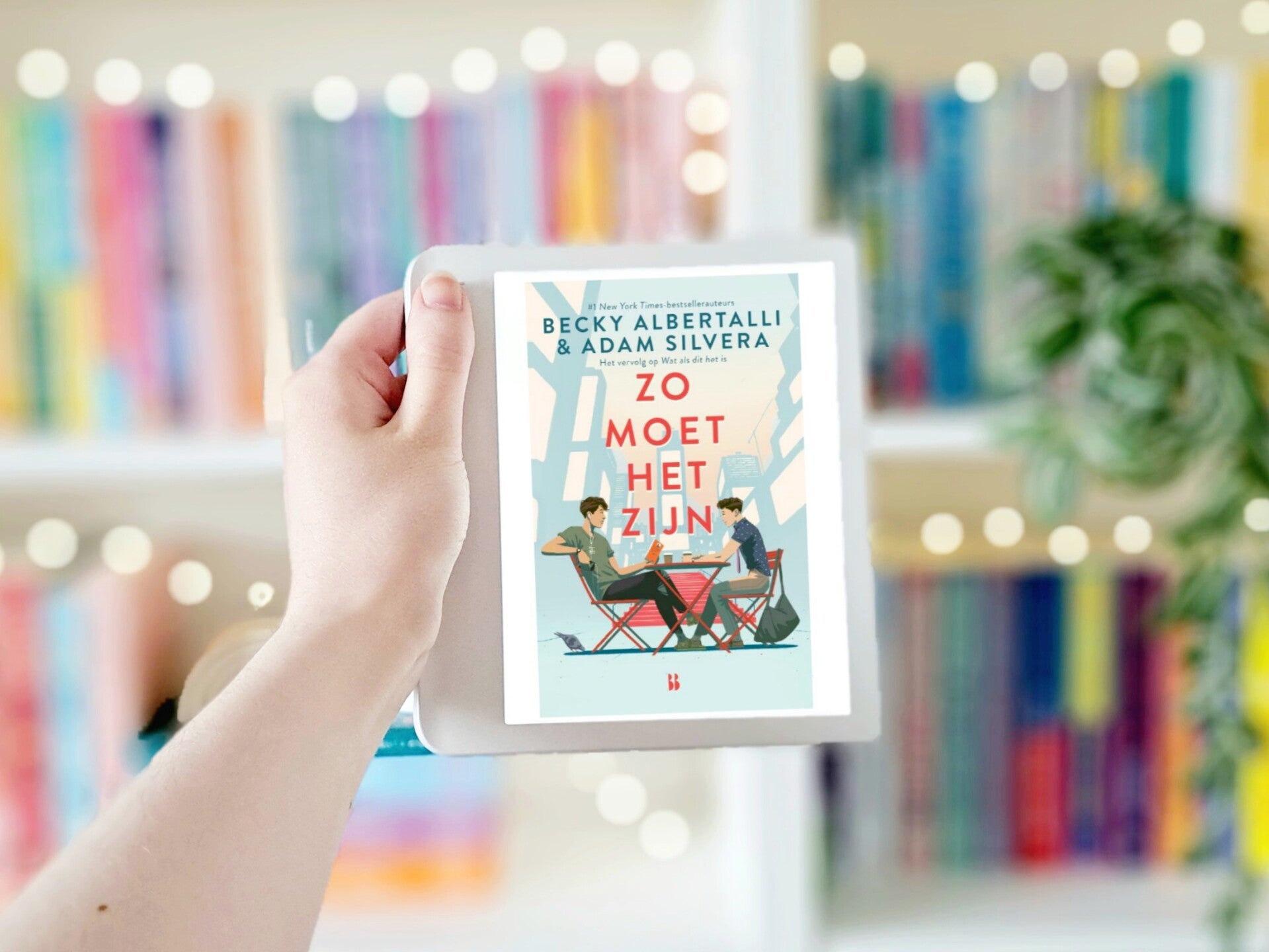 Een witte hand houdt een e-reader met de omslag van 'Zo moet het zijn' van Becky Albertalli en Adam SIlvera omhoog voor een kleurrijke wazige boekenkast met lichtjes. Recensie door turnthepages.nl | Disabled bookstagrammer en neurodivergente boekblogger.