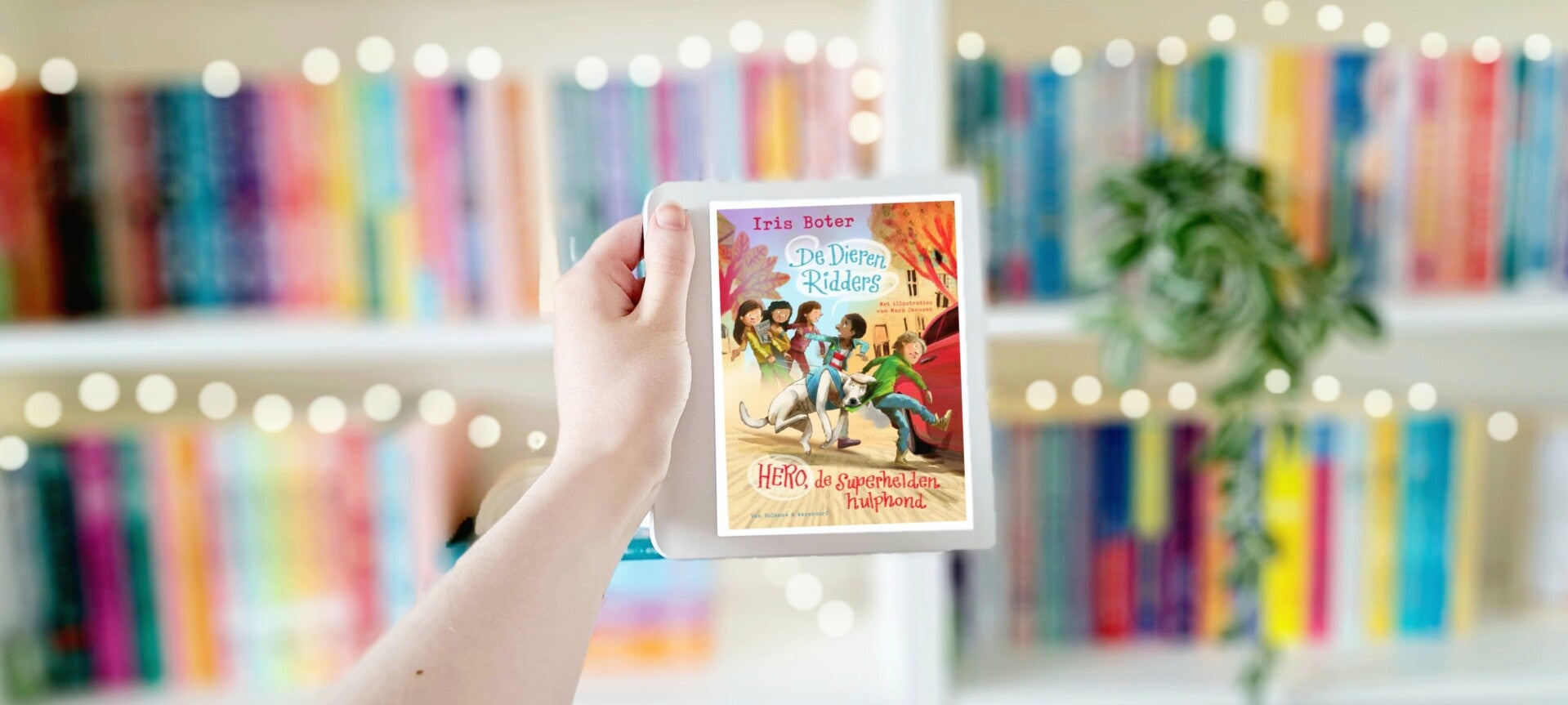 Een witte hand houdt een tablet met de omslag van De Dierenridders 1 'Hero, de superheldenhulphond' van Iris Boter en Mark Janssen omhoog voor een kleurrijke wazige boekenkast met lichtjes. Foto bij recensie door turnthepages.nl Disabeld bookstagrammer en