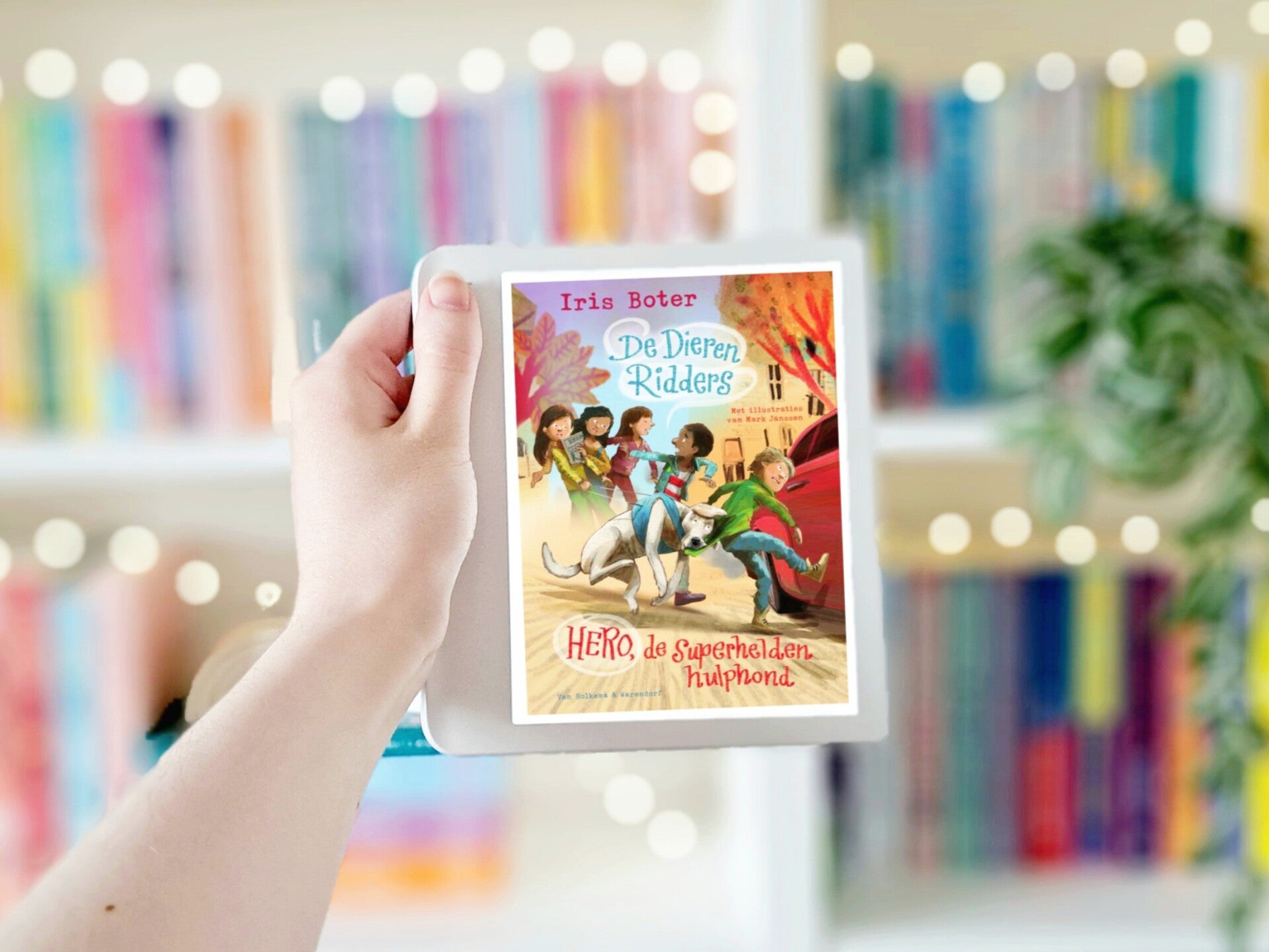 Een witte hand houdt een tablet met de omslag van De Dieren-Ridders deel 1: 'Hero, de superheldenhulphond' van Iris Boter en Mark Janssen omhoog voor een kleurrijke wazige boekenkast met lichtjes. Foto bij recensie door turnthepages.nl | Disabled bookstag
