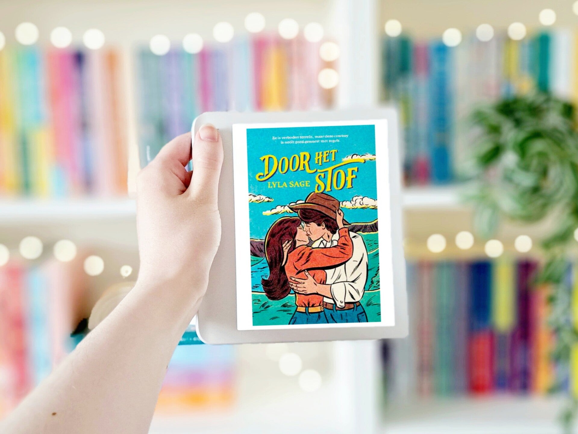 Een witte hand houdt 'Love redesigned' van Lauren Asher omhoog voor een kleurrijke wazige boekenkast met lichtjes. Foto bij recensie door turnthepages.nl | Disabled bookstagrammer en neurodivergente boekblogger.