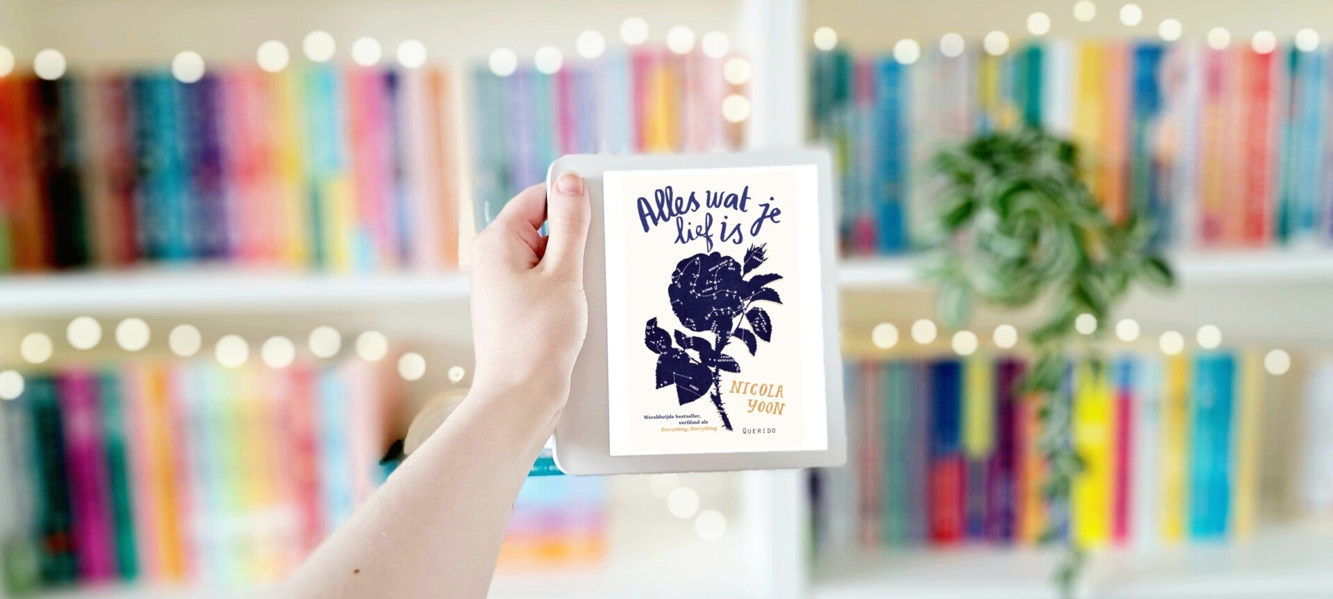 Een witte hand houdt 'Alles wat je lief is' van Nicola Yoon omhoog voor een kleurrijke wazige boekenkast met lichtjes. Foto bij recensie door turnthepages.nl  Disabled bookstagrammer en blogger.