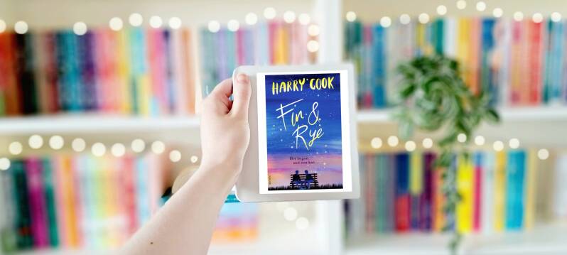 Een witte hand houdt 'Fin & Rye' van Harry Cook omhoog voor een kleurrijke wazige boekenkast met lichtjes. Foto bij recensie door turnthepages.nl Rolstoelgebruiker en disabled bookstagrammer en blogger.