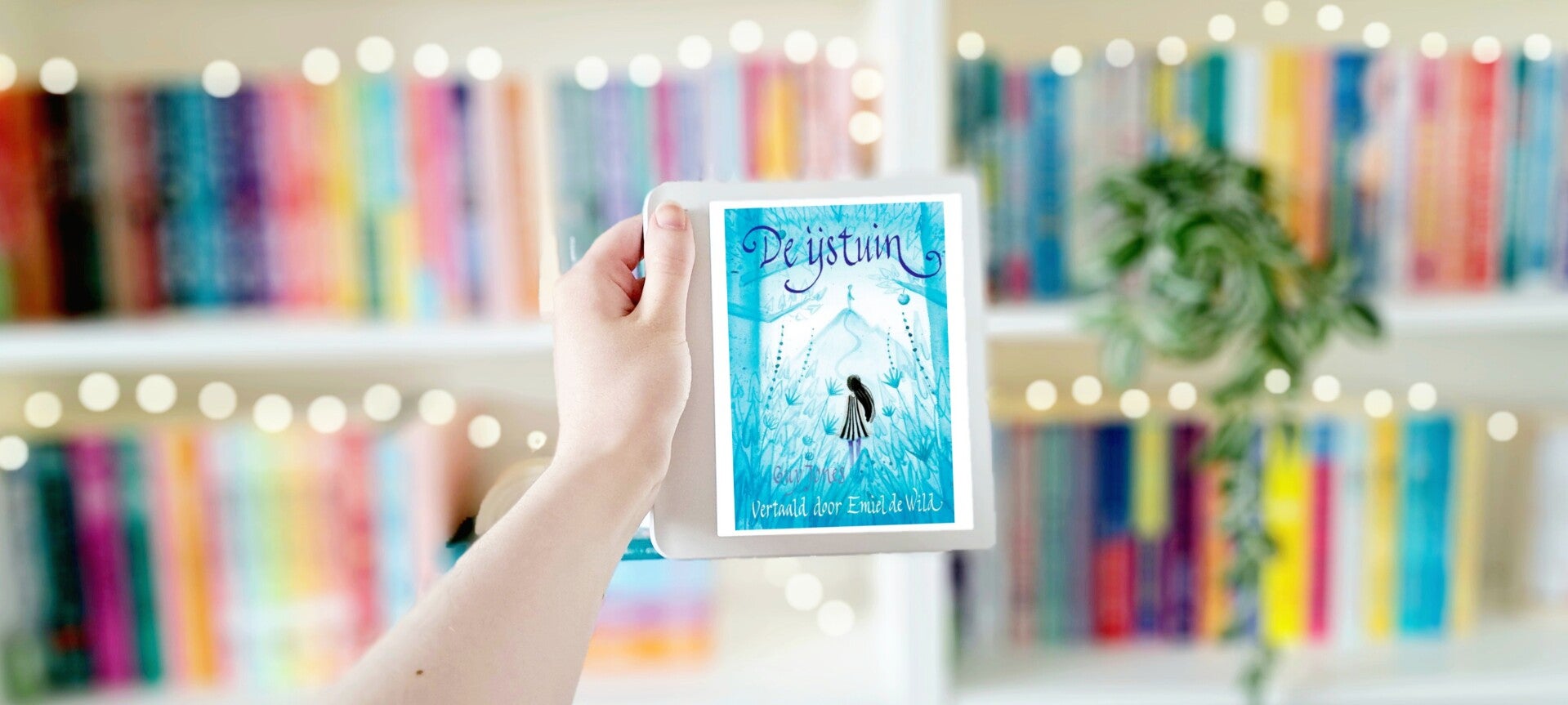 Een witte hand houdt 'De ijstuin' van Guy Jones omhoog tegen een wazige boekenkast met lichtjes. Foto bij recensie van turnthepages.nl Rolstoelgebruiker en disabled bookstagrammer en blogger.