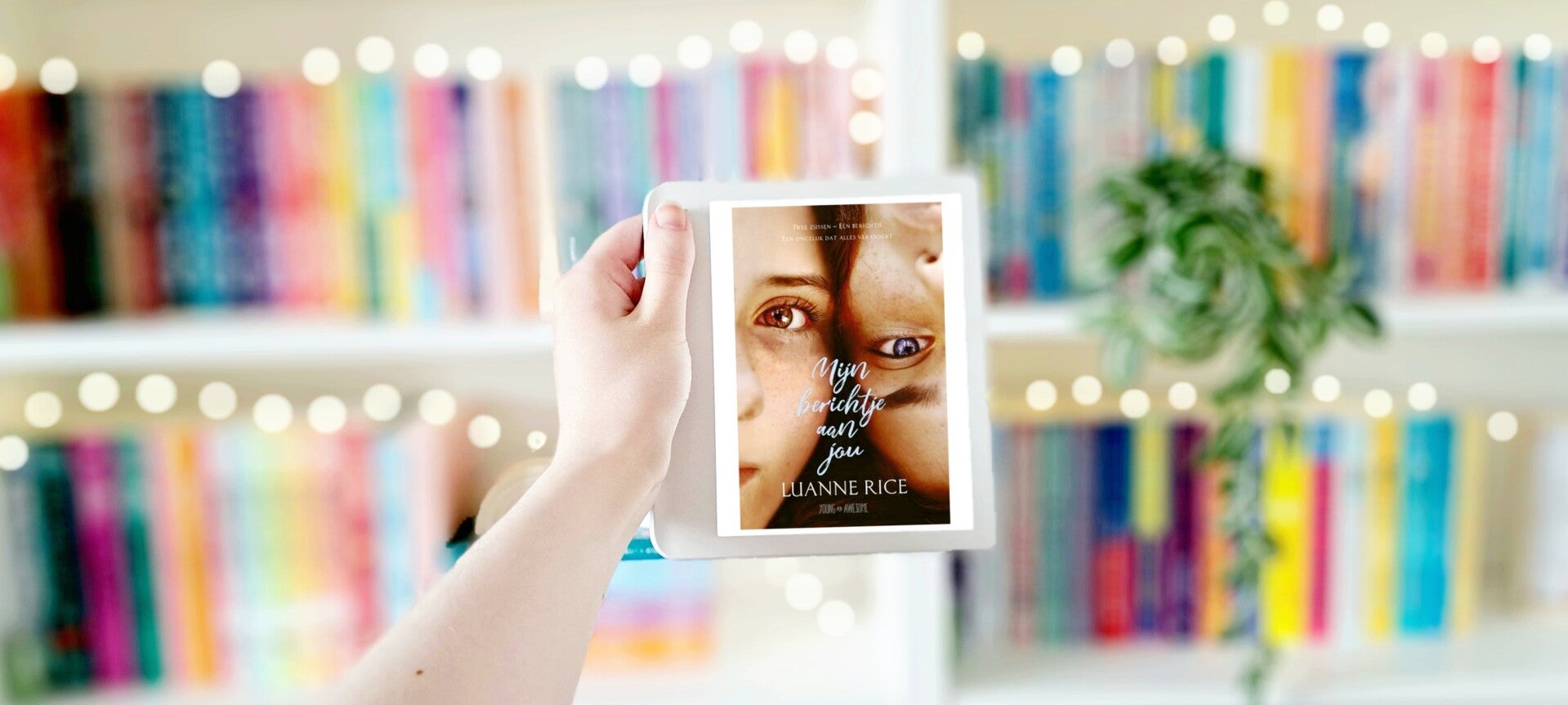 Een witte hand houdt 'Mijn berichtje aan jou' van Luanne Rice omhoog voor een kleurrijkew wazige boekenkast met lichtjes. Foto bij recensie door turnthepages.nl Rolstoelgebruiker en disabled bookstagrammer en blogger.