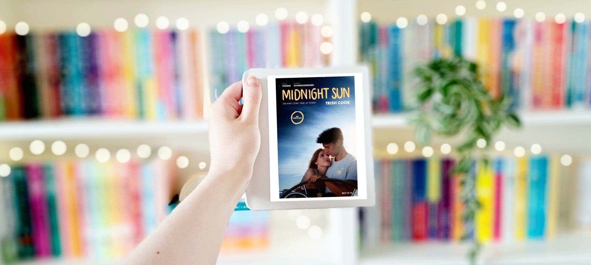 Een witte hand houdt 'Mignight Sun' van Trish Cook omgoof voor een kleurrijke wazige boekenkast met lichtjes. Foto bij recensie door turnthepages.nl Disabled bookstagrammer en blogger.
