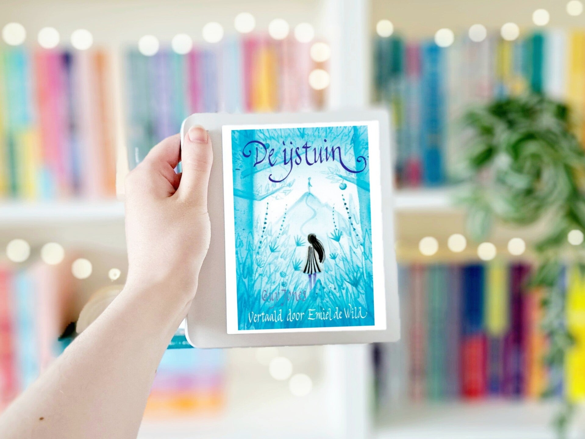 Een witte hand houdt 'De ijstuin' van Guy Jones omhoog tegen een wazige boekenkast met lichtjes. Foto bij recensie van turnthepages.nl Rolstoelgebruiker en disabled bookstagrammer en blogger.