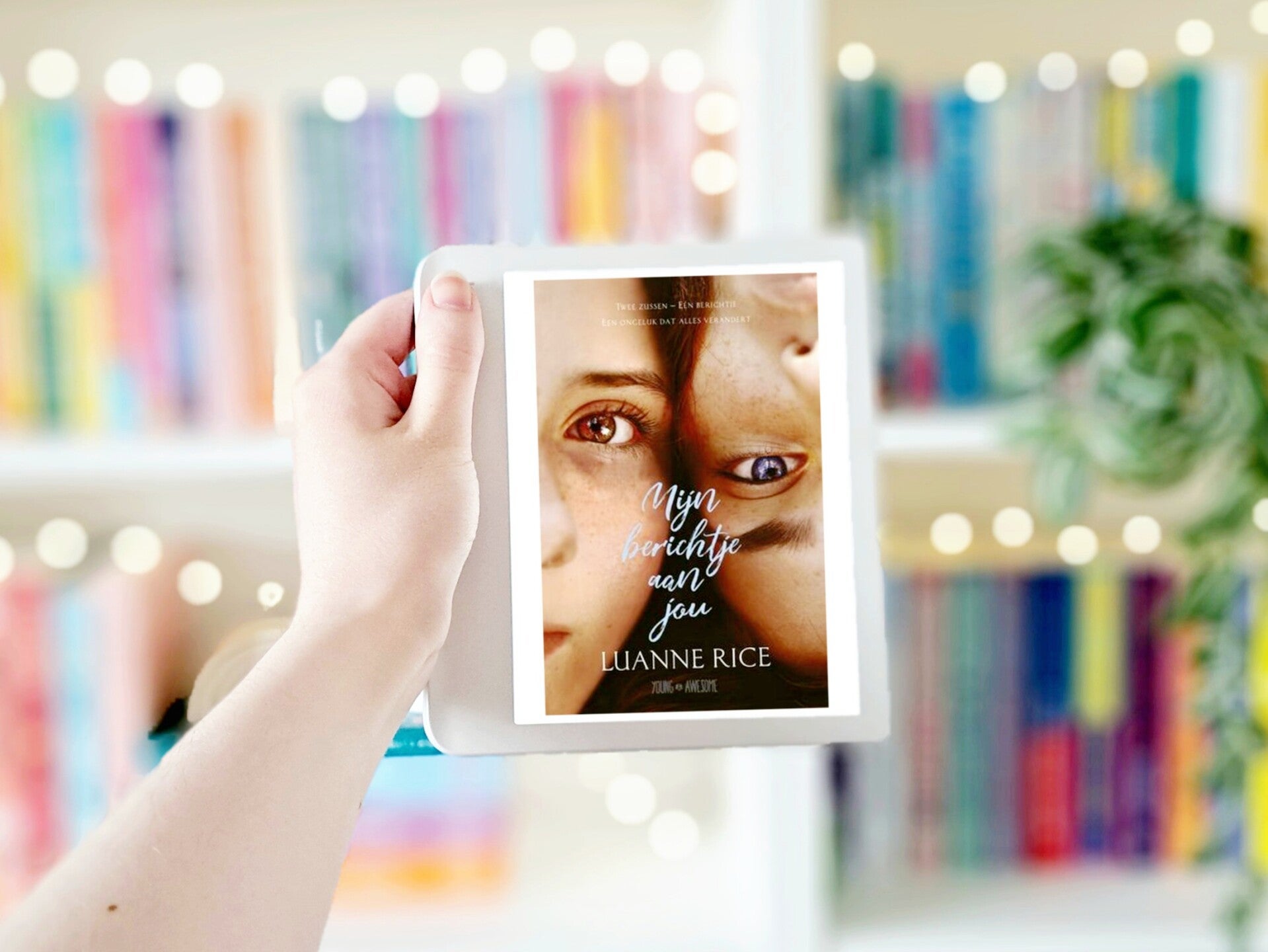 Een witte hand houdt 'Mijn berichtje aan jou' van Luanne Rice omhoog voor een kleurrijke wazige boekenkast met lichtjes.  Foto bij recensie door turnthepages.nl Rolstoelgebruiker en disabled bookstagrammer en blogger.