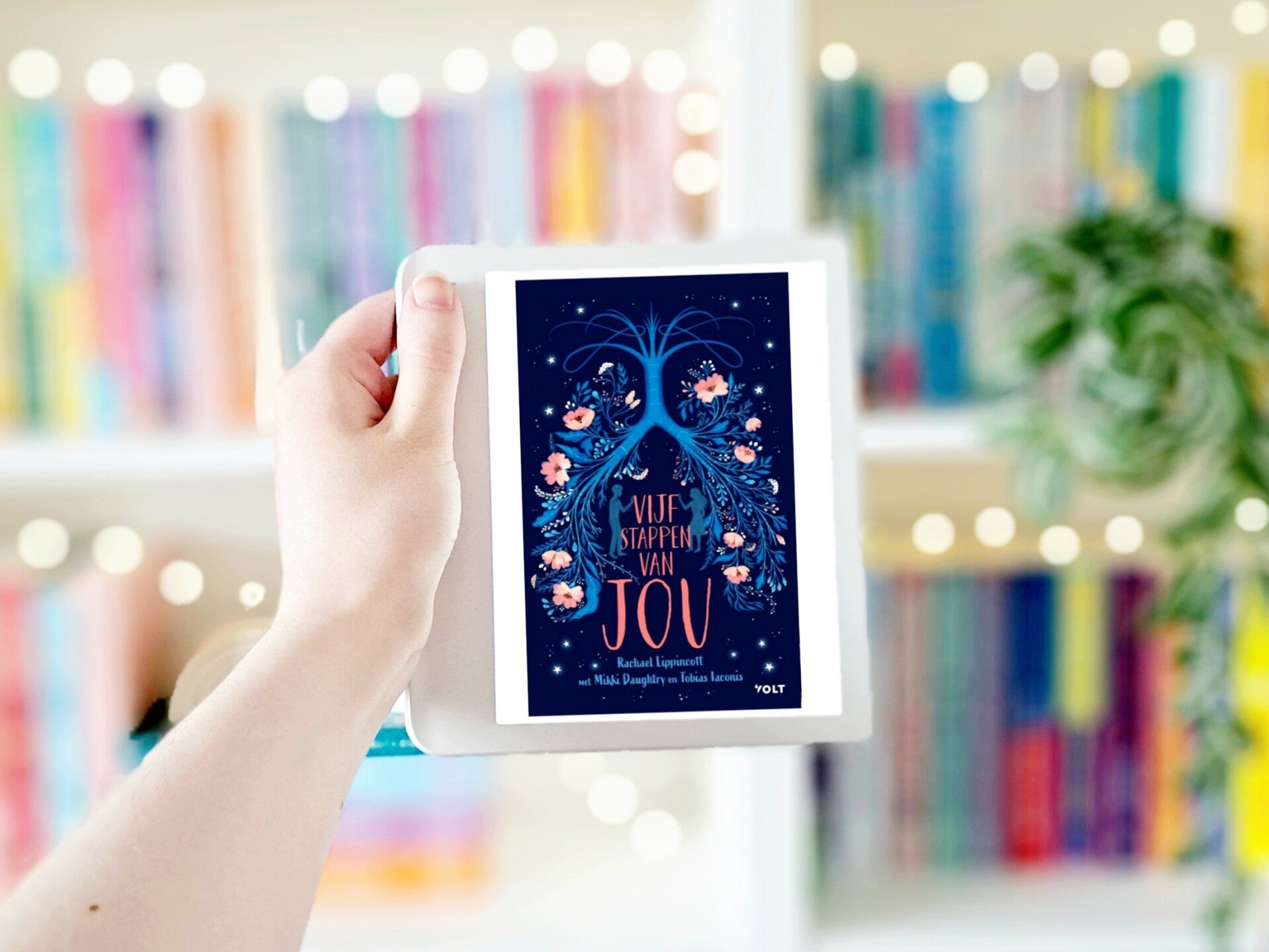 Een witte hand houdt 'Vijf stappen van jou' van Rachael Lippincott, Mikki Daughtry en Tobias Iaconis omhoog voor een kleurrijke wazige boekenkast met lichtjes. Foto bij recensie door turnthepages.nl Disabled bookstagrammer en blogger.