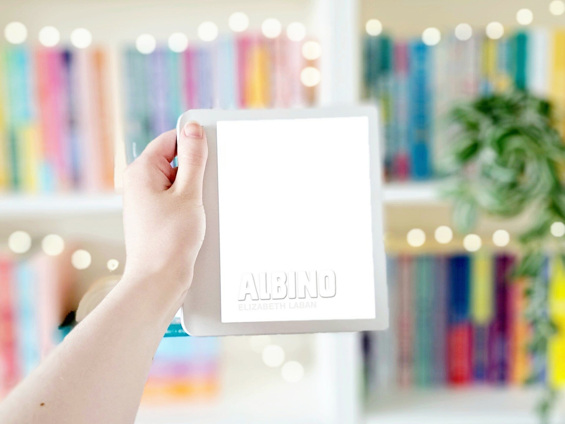 Een witte hand houdt 'Albino' van Elizabeth LaBan omhoog voor een kleurrijke wazige boekenkast met lichtjes. Foto bij recensie door turnthepages.nl Rolstoelgebruiker, disabled bookstagrammer en queer blogger