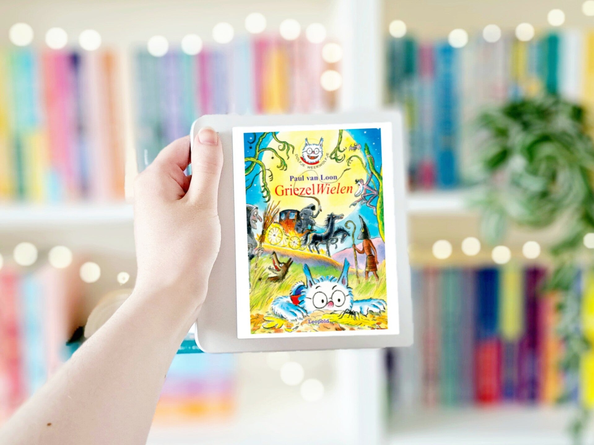 Een witte hand houdt 'GriezelWielen' van Paul van Loon omhoog voor een kleurrijke wazige boekenkast met lichtjes. Foto bij own-voices recensie door turnthepages.nl Rolstoelgebruiker en disabled bookstagrammer en blogger.