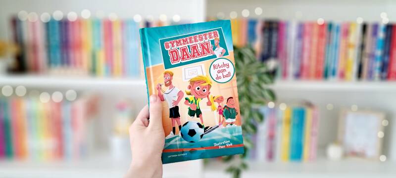 Een witte hand houdt 'Gymmeester Daan 1: Ritchy aan de bal!' omhoog voor een kleurrijke wazige boekenkast met lichtjes. Recensie door turnthepages.nl | Disabled bookstagrammer, chronisch zieke rolstoelgebruiker en neurodivergente boekblogger met ADHD.
