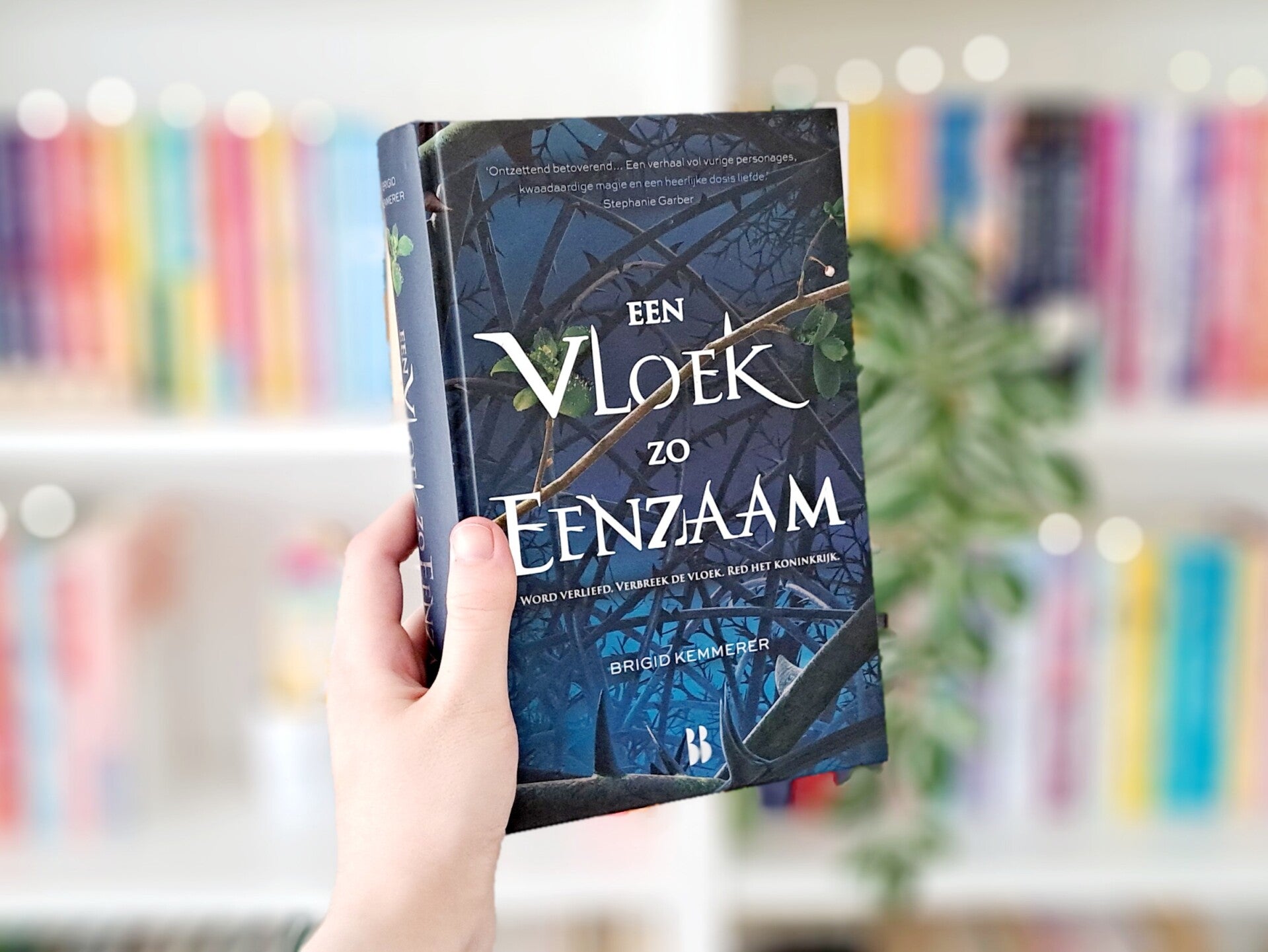 Een witte hand houdt 'Een vloek zo eenzaam' van Brigid Kemmerer omhoog voor een kleurrijke wazige boekenkast met lichtjes. Foto bij recensie door turnthepages.nl Rolstoelgebruiker en disabled bookstagrammer en blogger.