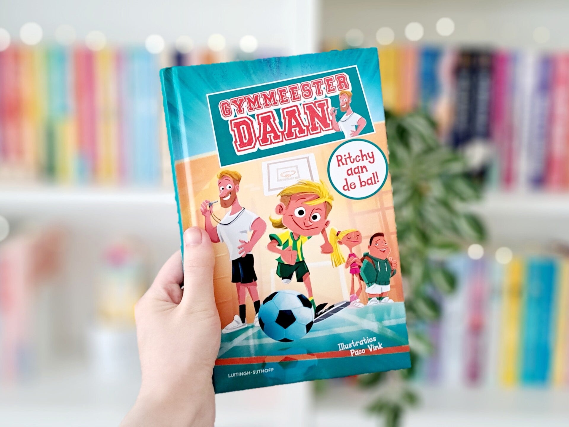 Een witte hand houdt 'Recept voor een regelrechte ramp' van Linda van de Pol omhoog voor een kleurrijke wazige boekenkast met lichjtes. Foto bij recensie door turnthepages.nl Rolstoelgebruiker en disabled bookstagrammer en blogger met ADHD.