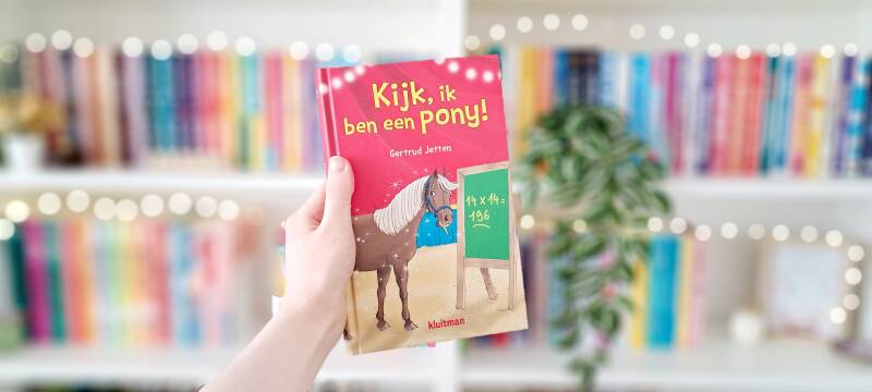 Een witte hand houdt 'Kijk, ik ben een pony!' van Gertrud Jetten omhoog voor een kleurrijke wazige boekenkast met lichtjes. | Recensie door turnthepages.nl | Rolstoelgebruiker, disabled bookstagrammer en chronisch zieke boekblogger met ADHD.
