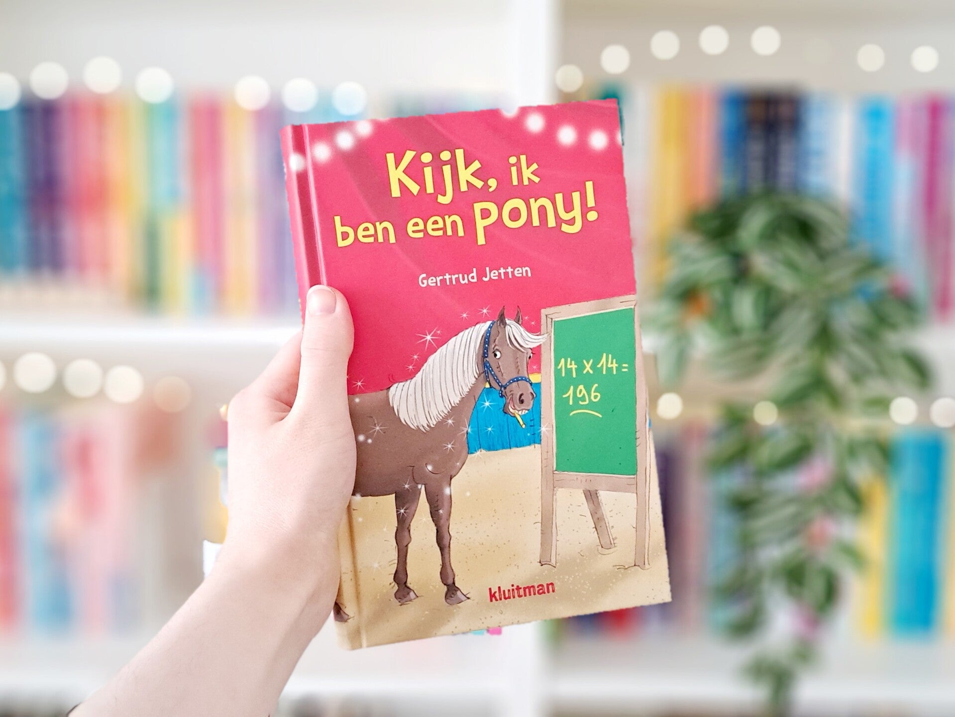 Een witte hand houdt 'Kijk, ik ben een pony!' van Gertrud Jetten omhoog voor een kleurrijke wazige boekenkast met lichtjes. Recensie door turnthepages.nl | Disabled bookstagrammer, chronisch zieke rolstoelgebruiker en neurodivergent boekblogger.