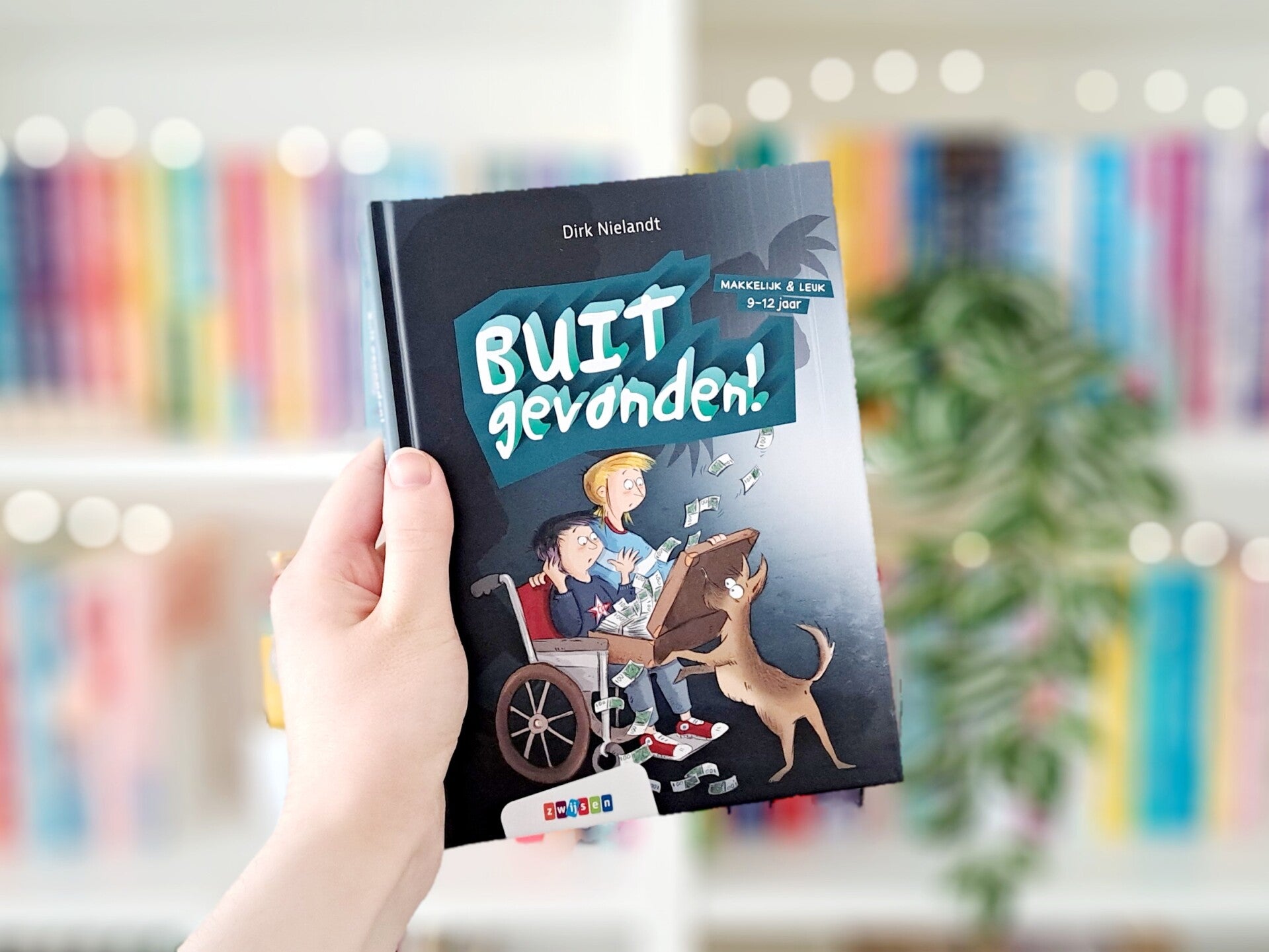 Een witte hand houdt 'Buit gevonden!' van Dirk Nielandt omhoog voor een kleurrijke wazige boekenkast met lichtejs. | Recensie door turnthepages.nl | Rolstoelgebruiker, disabled bookstagrammer en chronisch zieke boekblogger met ADHD.