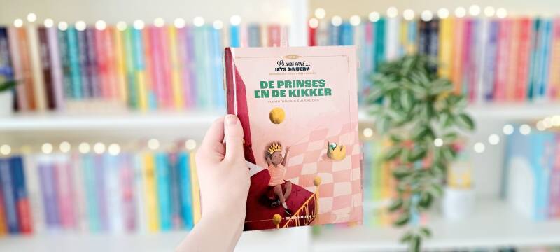 Een witte hand houdt 'De prinses en de kikker' van Floor Tinga en Evi Radoes omhoog voor een kleurrijke wazige boekenkast met lichtjes. Recensie door Annemarie van turnthepages.nl