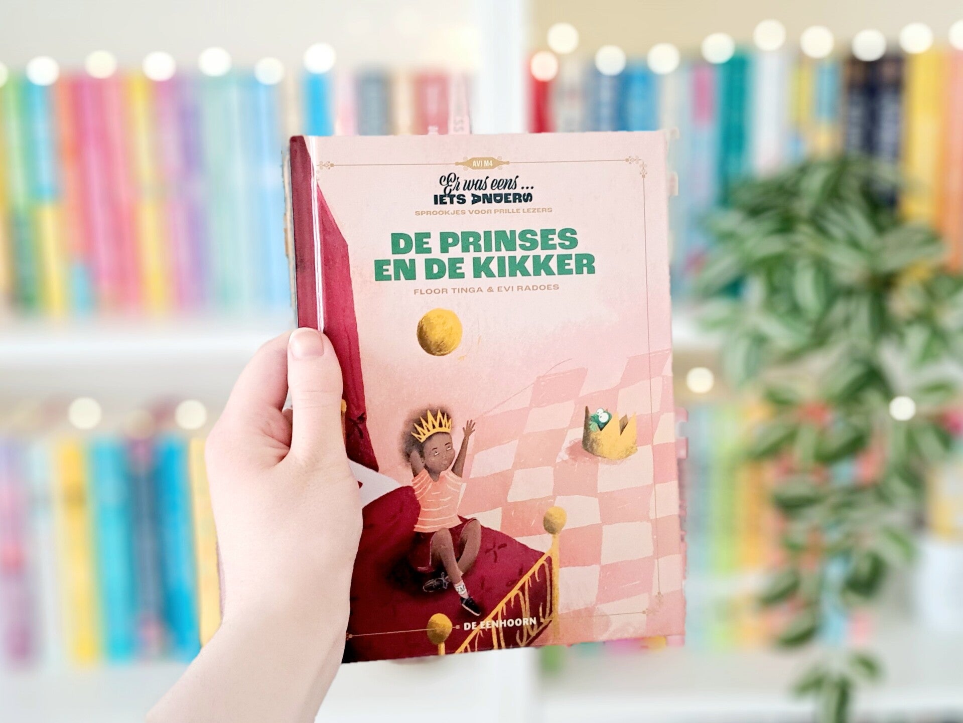 Een witte hand houdt 'De prinses en de kikker' van Floor Tinga en Evi Radoes omhoog voor een kleurrijke wazige boekenkast met lichtjes. Recensie door Annemarie van turnthepages.nl