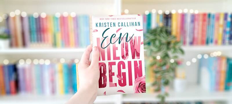 Een witte hand houdt 'een nieuw begin' van Kristen Callihan omhoog voor een kleurrijke wazige boekenkast met lichtjes. own-voices recensie door Annemarie van turnthepages.nl | Disabled en neurodivergent boekblogger en boekstagrammer.