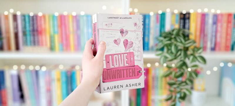 Een witte hand houdt 'love unwritten' van Lauren Asher omhoog voor een kleurrijke wazige boekenkast met lichtjes. Recensie door Annemarie van turnthepages.nl | Disabled en neurodivergent boekblogger en bookstagrammer.
