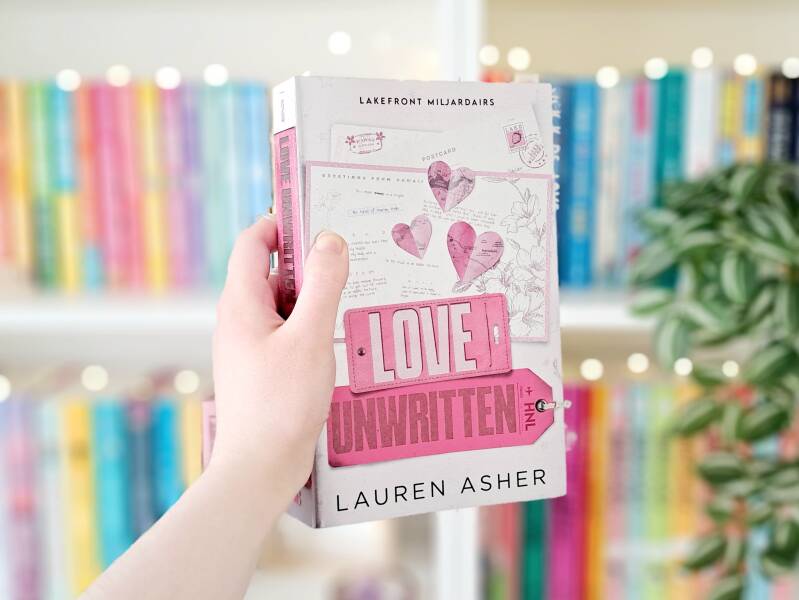 Een witte hand houdt 'love unwritten' van Lauren Asher omhoog voor een kleurrijke wazige boekenkast met lichtjes. Recensie door Annemarie van turnthepages.nl | Disabled en neurodivergent boekblogger en bookstagrammer