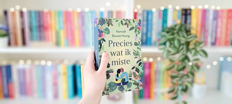 Een witte hand houdt 'precies wat ik miste' van Hannah Bonam-Young omhoog voor een kleurrijke wazige boekenkast met lichtjes. Recensie door Annemarie van turnthepages.nl