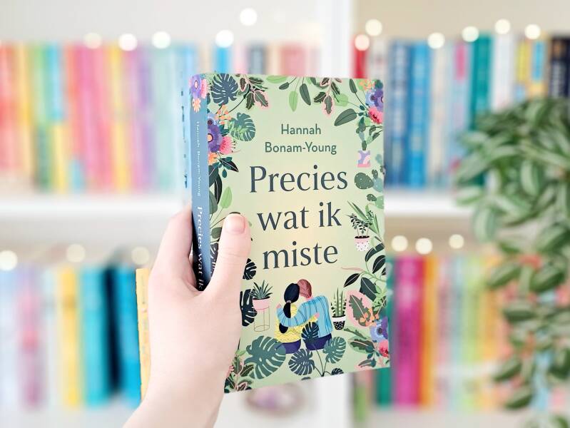 Een witte hand houdt 'precies wat ik miste' van Hannah Bonam-Young ohoog voor een kleurrijke wazige boekenkast met lichtjes. Recensie door Annemarie van turnthepages.nl | Disabled bookstagrammer en boekblogger.